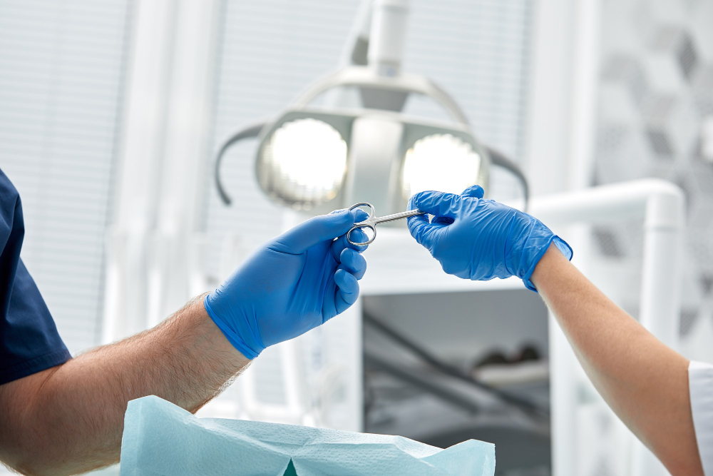 Zbliżenie na dłonie dwóch osób w niebieskich rękawiczkach chirurgicznych, podających sobie metalowe nożyczki chirurgiczne, na tle jasnych lamp zabiegowych.